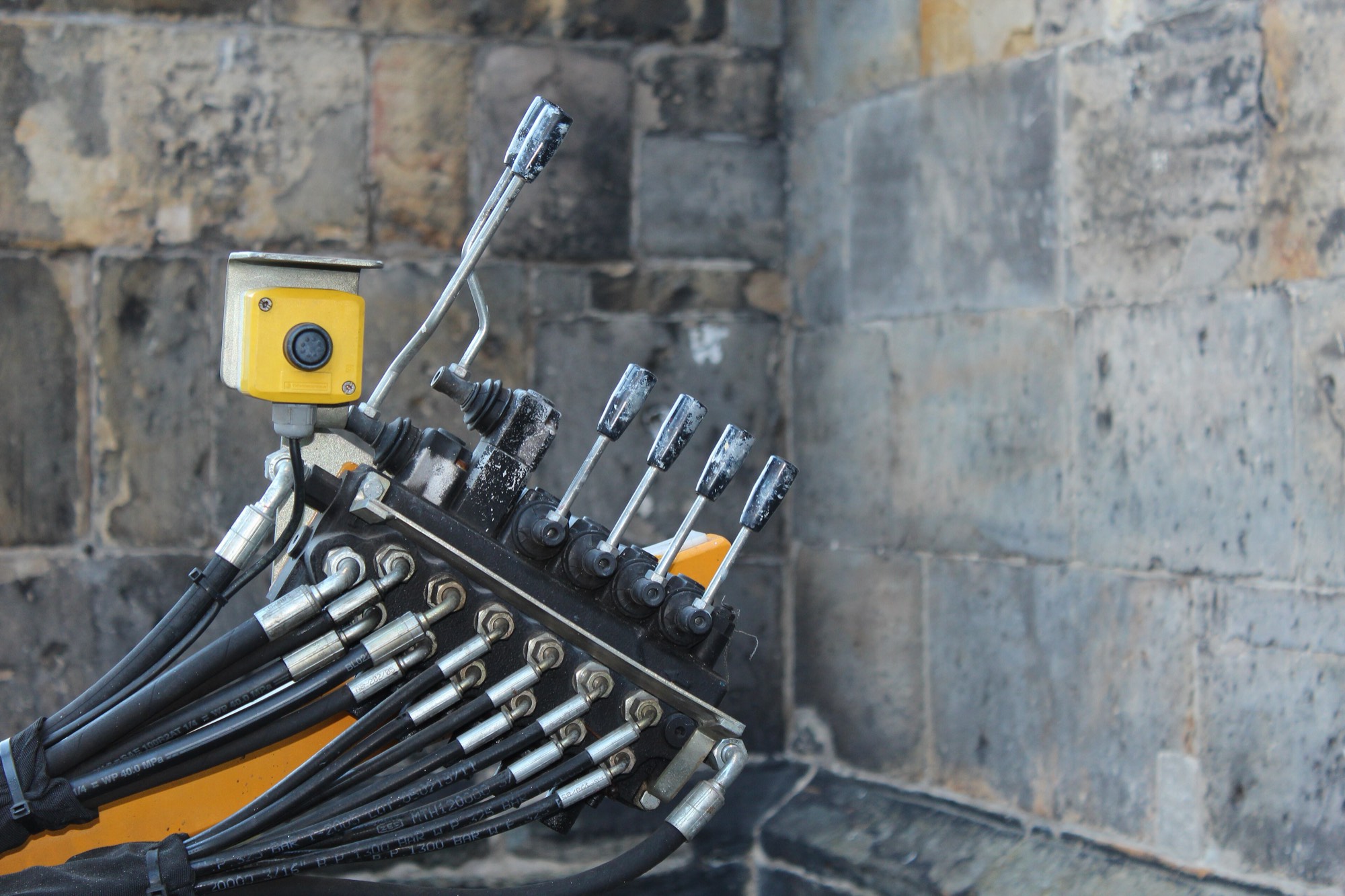 A bank of hydraulic control valves against a stone wall, spool levers above, hose fittings below